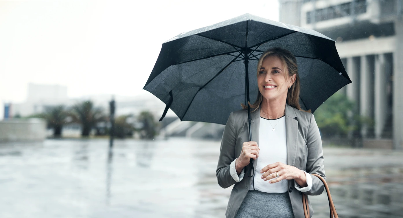 Smiling woman in business attire holding an umbrella in the rain, symbolising resilience and financial protection during uncertain times.