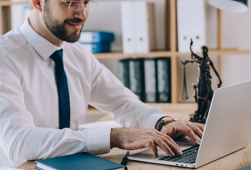 Legal professional in a shirt and tie working on a laptop at his office desk, with legal books and Lady Justice statue in the background.